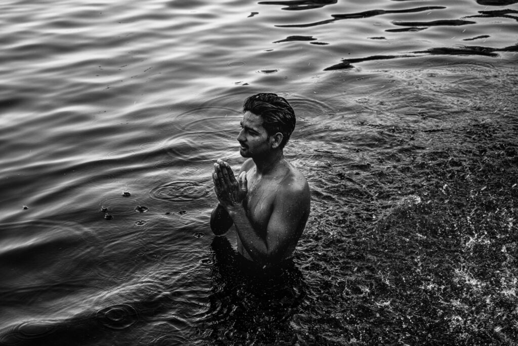 Shirtless man praying in river waters at Varanasi, India, captured in black and white.