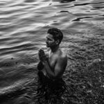 Shirtless man praying in river waters at Varanasi, India, captured in black and white.