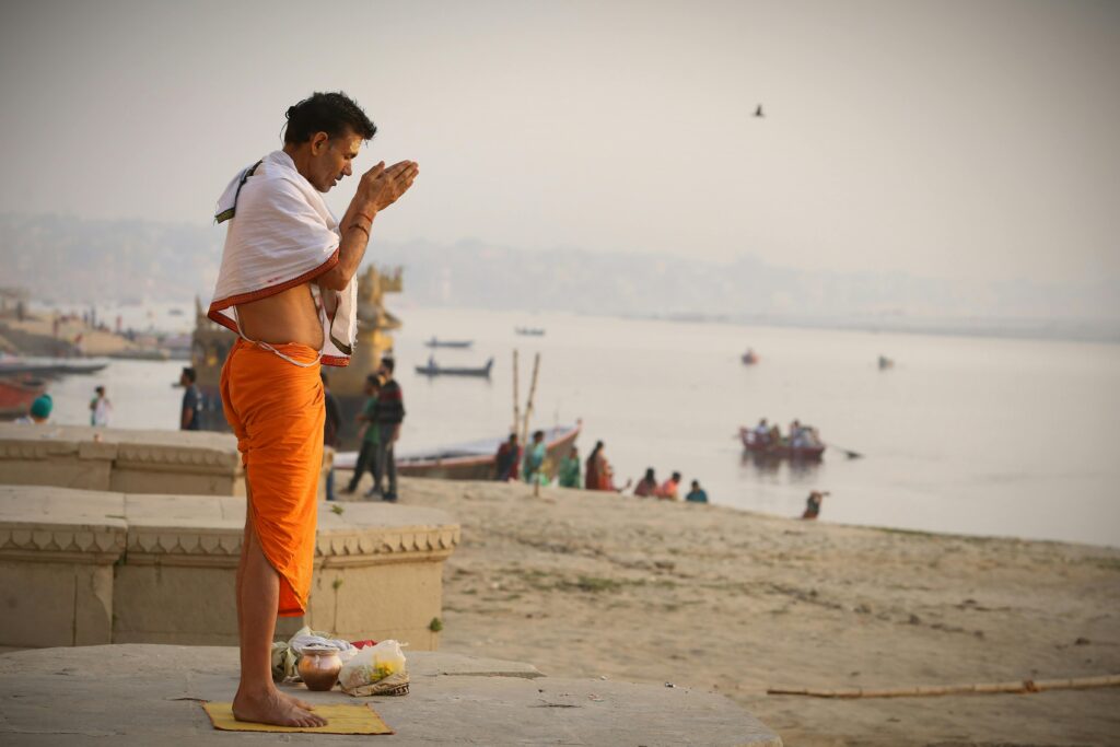 Devout man in orange robe praying on the riverbank at sunrise, a serene and spiritual moment.